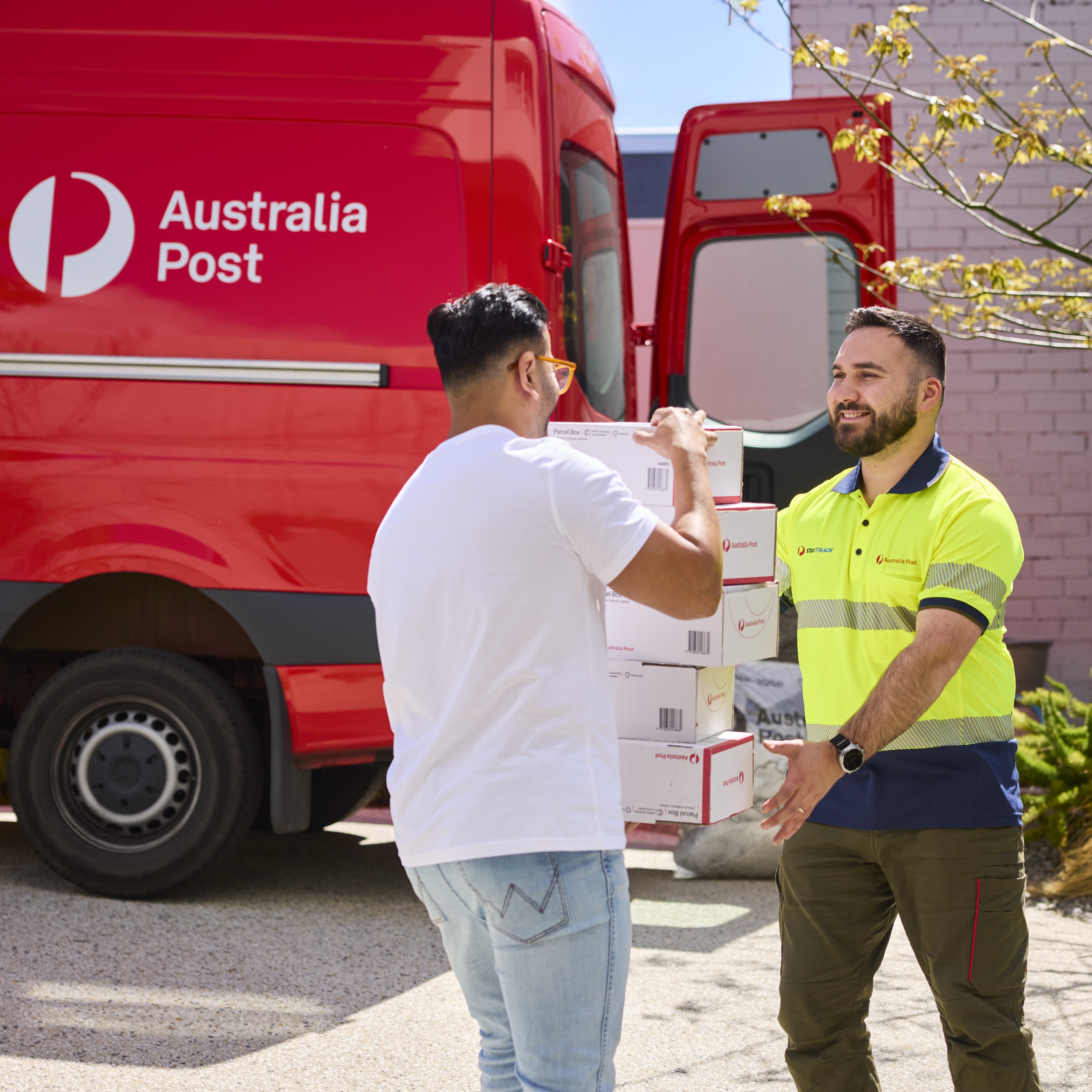 Two people transferring parcel boxes beside a red Parcel and Send van with its rear door open.